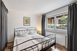 Ein Einzelbett mit Metallgestell, ein Nachttisch mit Lampe, ein gerahmtes Bild an der Wand und ein Fenster mit Vorhängen.