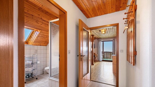 A hallway with wooden ceilings, a visible bathroom with a toilet and towel rack, and a view of an outdoor deck.