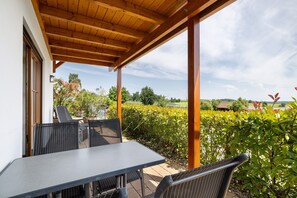 Eine überdachte Terrasse mit Tisch und Stühlen, einer Holzbalkendecke und Blick auf einen Weinberg sowie entfernte Gebäude.