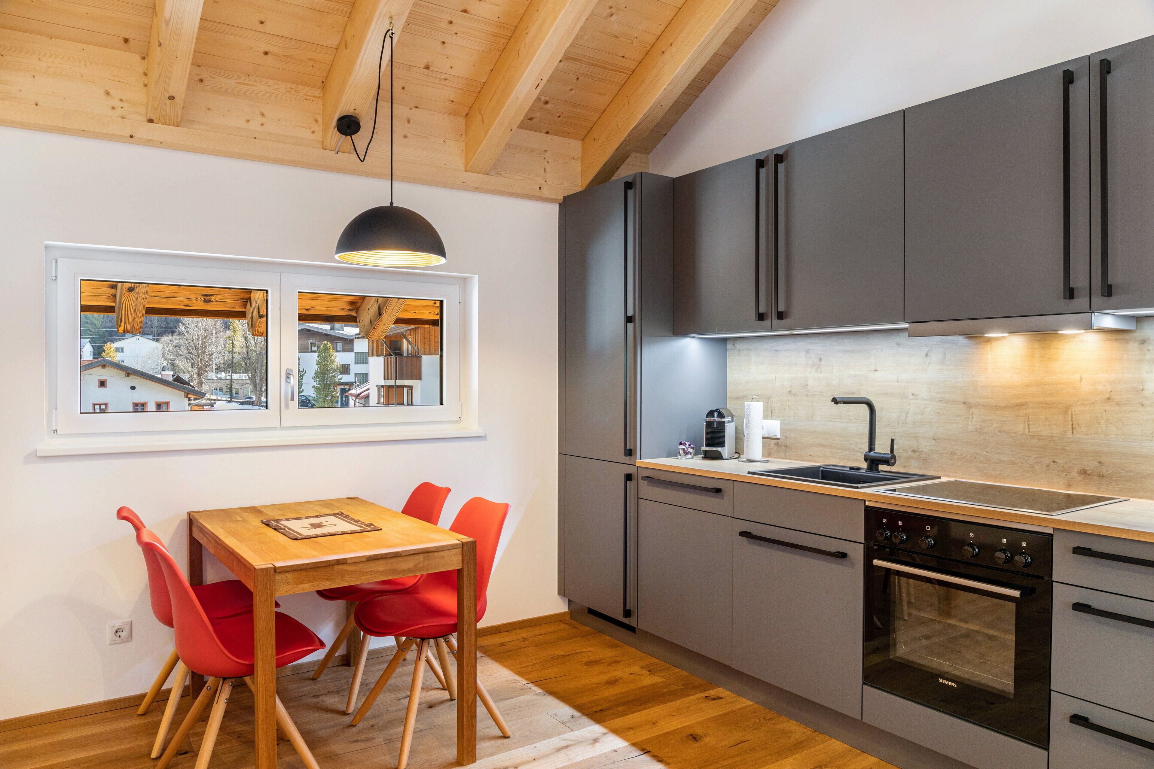 A modern kitchen with grey cabinets, a wooden dining table with red chairs, and a view through the window.