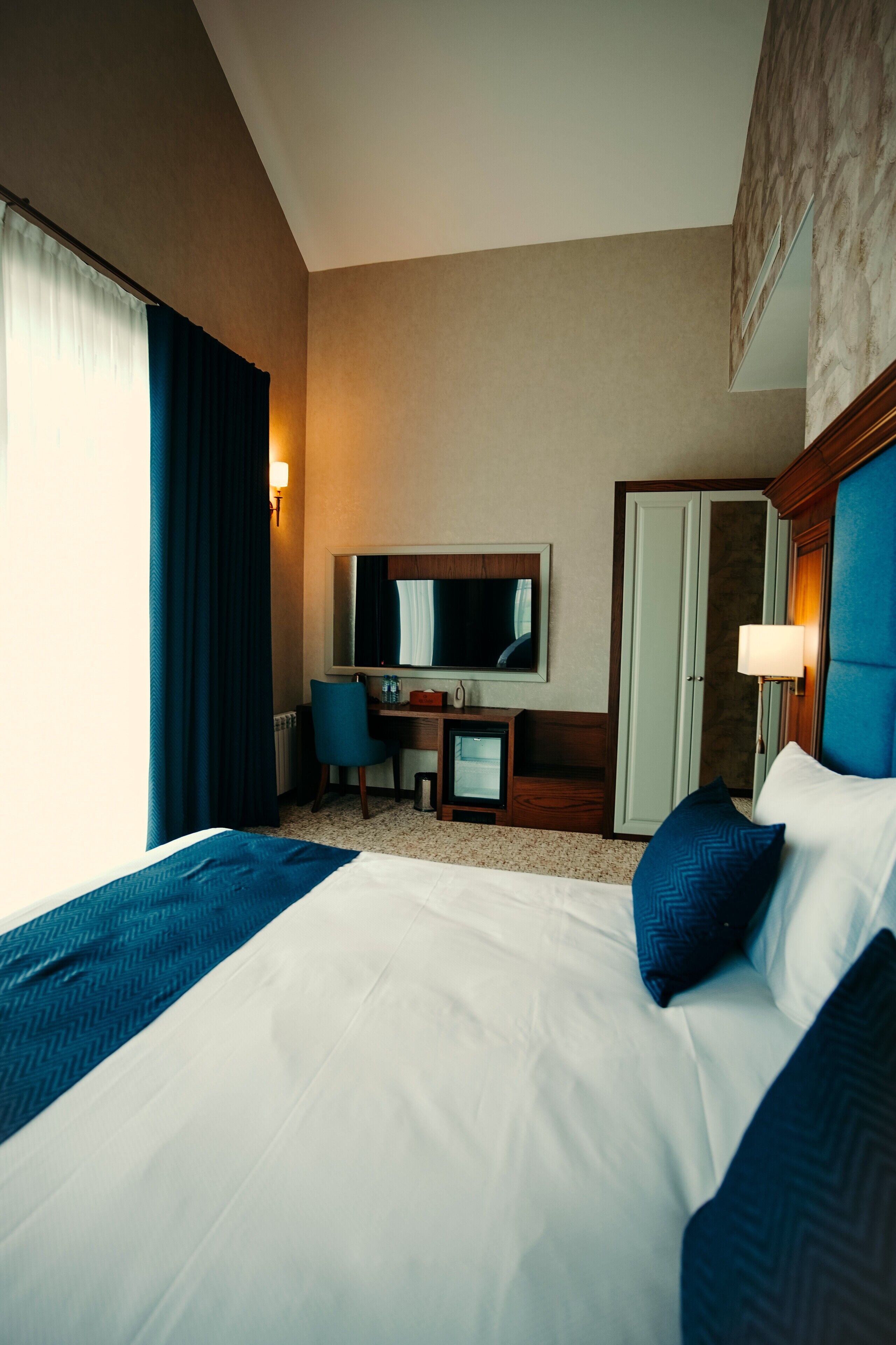 A hotel room with a large bed, a desk, a chair, a television, and a wardrobe.