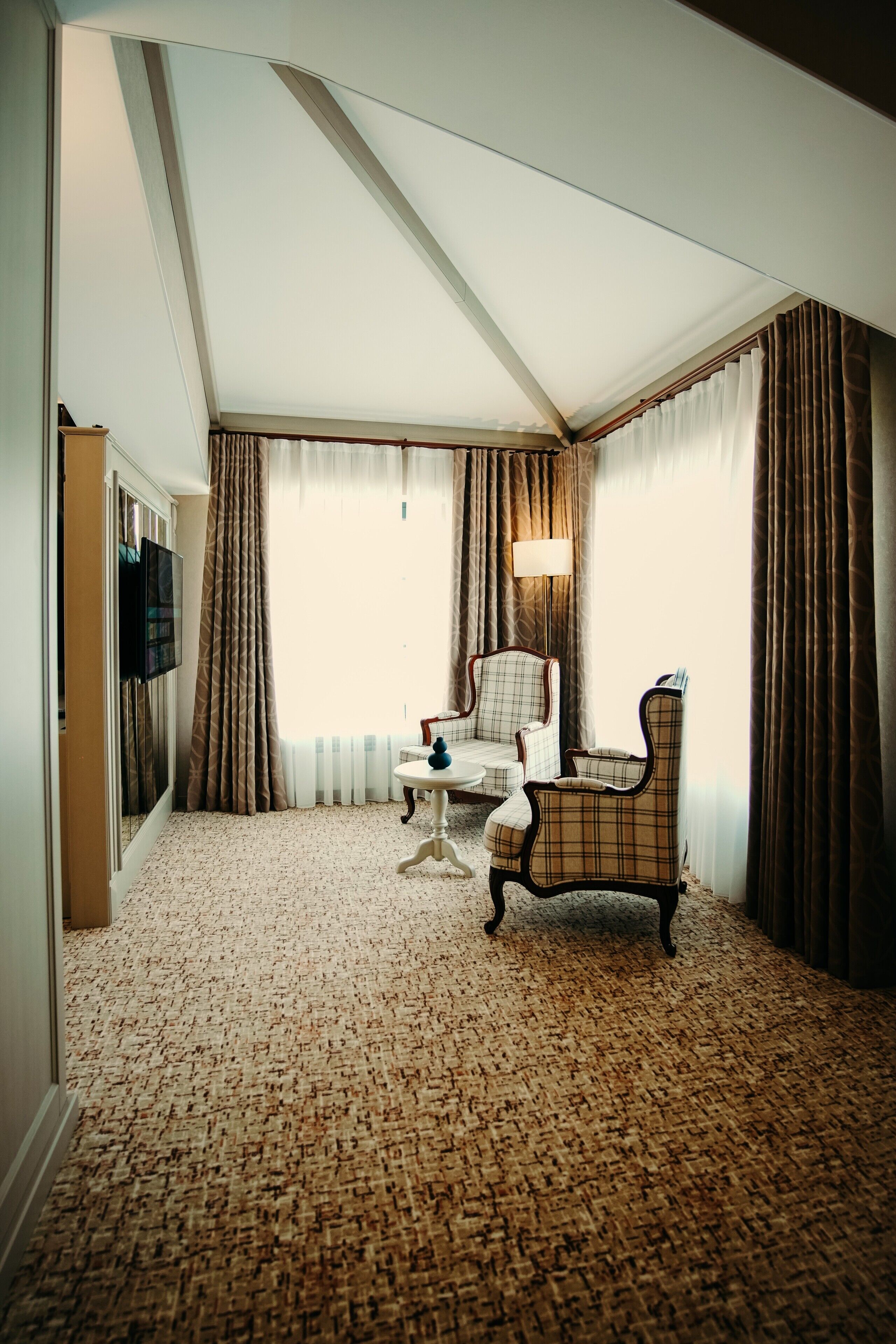 A hotel room with a checkered armchair, a small round table, a television mounted on the wall, and a large window with curtains.