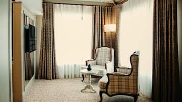 A hotel room with a checkered armchair, a small round table, a television mounted on the wall, and a large window with curtains.