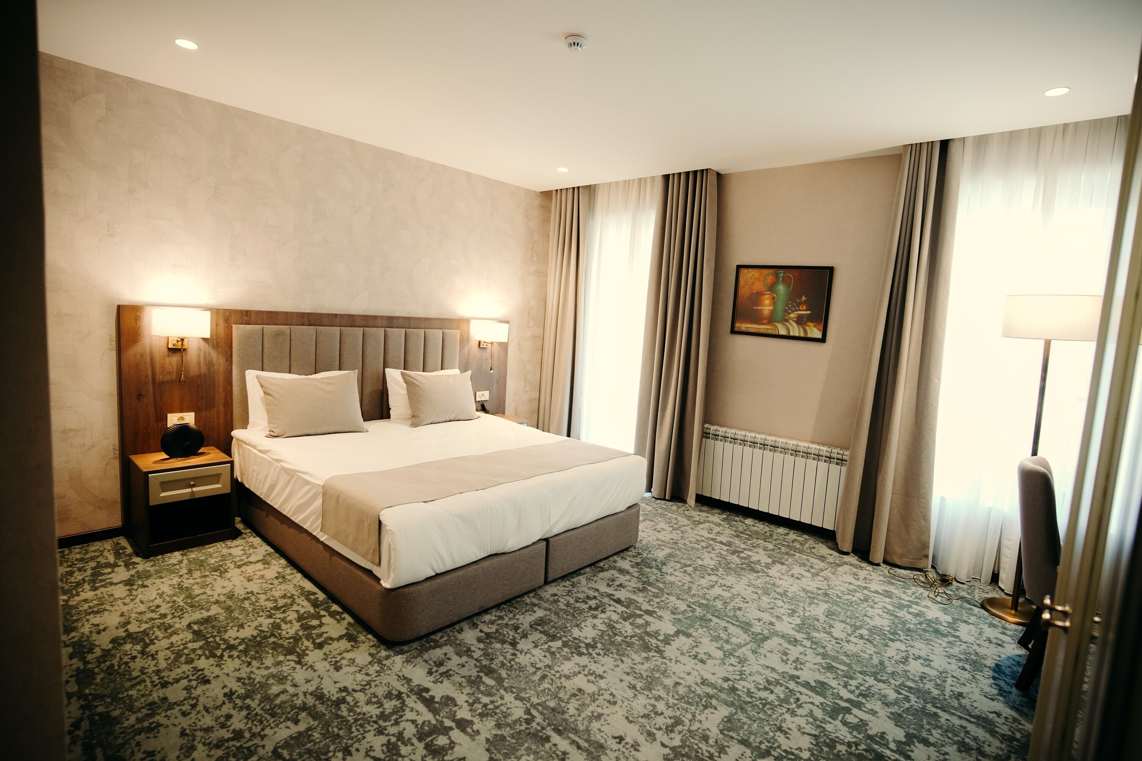 Una habitación de hotel con una cama grande, dos mesitas de noche con lámparas, un radiador y un cuadro en la pared.