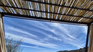 Un espace extérieur aménagé avec des sièges, offrant une vue sur un champ verdoyant et des collines. On y trouve une chaise en osier, une table avec de la nourriture et un verre, ainsi qu'un livre intitulé « Magisch Piemonte ».