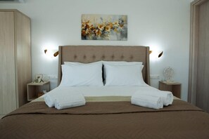 A neatly made bed with white linens and a brown bedspread, flanked by two bedside tables with decorative items, in a room with a neutral color scheme and a painting on the wall.