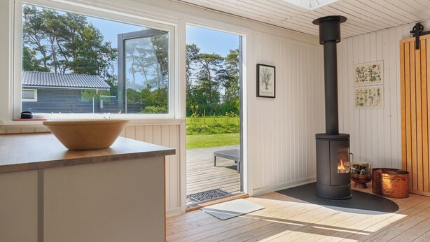 A modern interior with a large window, a wooden deck outside, and a wood-burning stove.