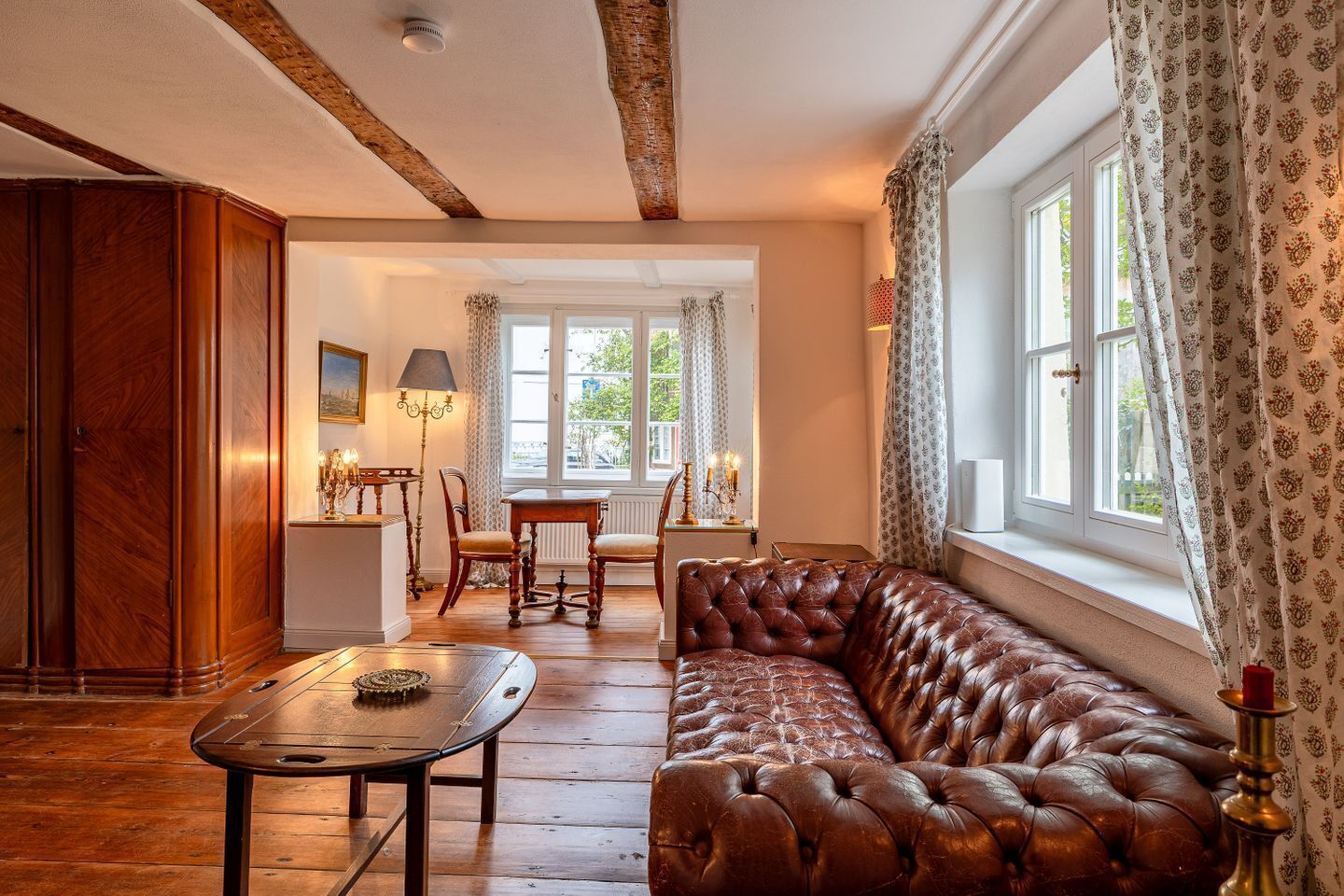 A room with a leather sofa, wooden table, and chairs. There is a window with patterned curtains and a view of greenery outside.