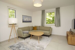 A modern living room with a beige sofa, a round wooden coffee table, and a large window with curtains. - Beachfront vacation rental with a terrace, access to the pool and sauna - Rex Rugia - Rex (Göhren)