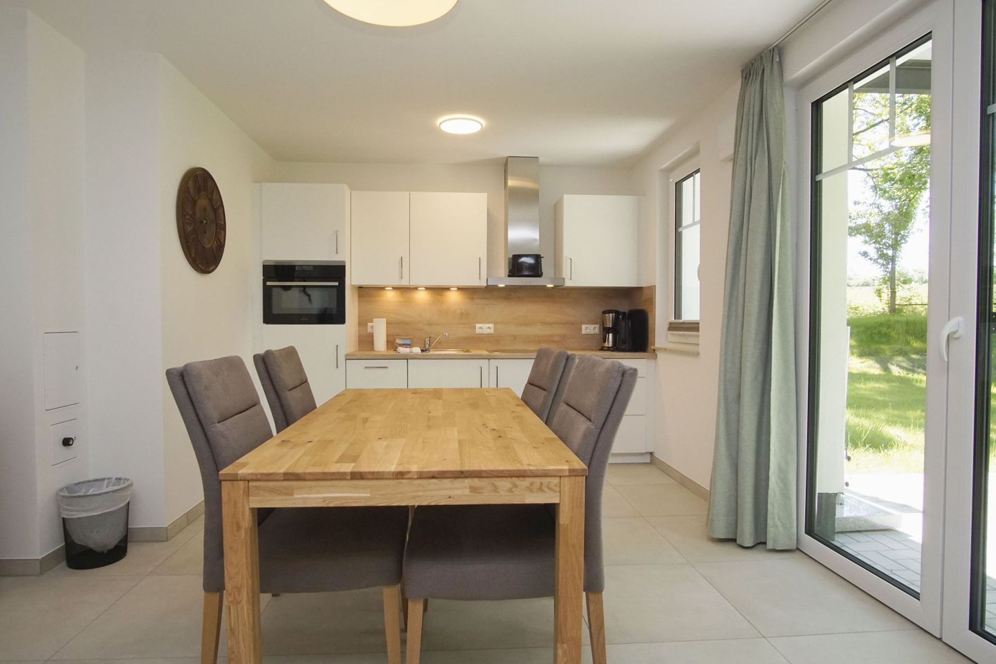 Une cuisine moderne avec des meubles blancs, une table à manger en bois et une vue sur un paysage verdoyant, visible à travers la porte-fenêtre coulissante.