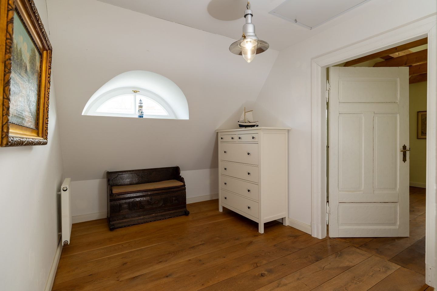 Ein Zimmer mit Holzboden, einer Bank, einem Schminktisch, einer Tür und einem Fenster mit Oberlicht.