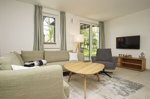 A modern living room with a sofa, armchair, wooden coffee table, and a TV on a stand. - Beachfront vacation rental with a terrace, access to the pool and sauna - Rex Rugia - Rex (Göhren)