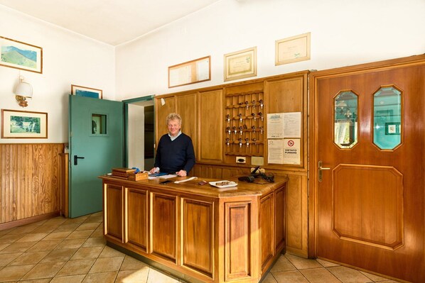 A reception desk with a wooden finish, a wall-mounted key rack, framed certificates, and a door with a glass panel. - albergo capannette di pei (Zerba)