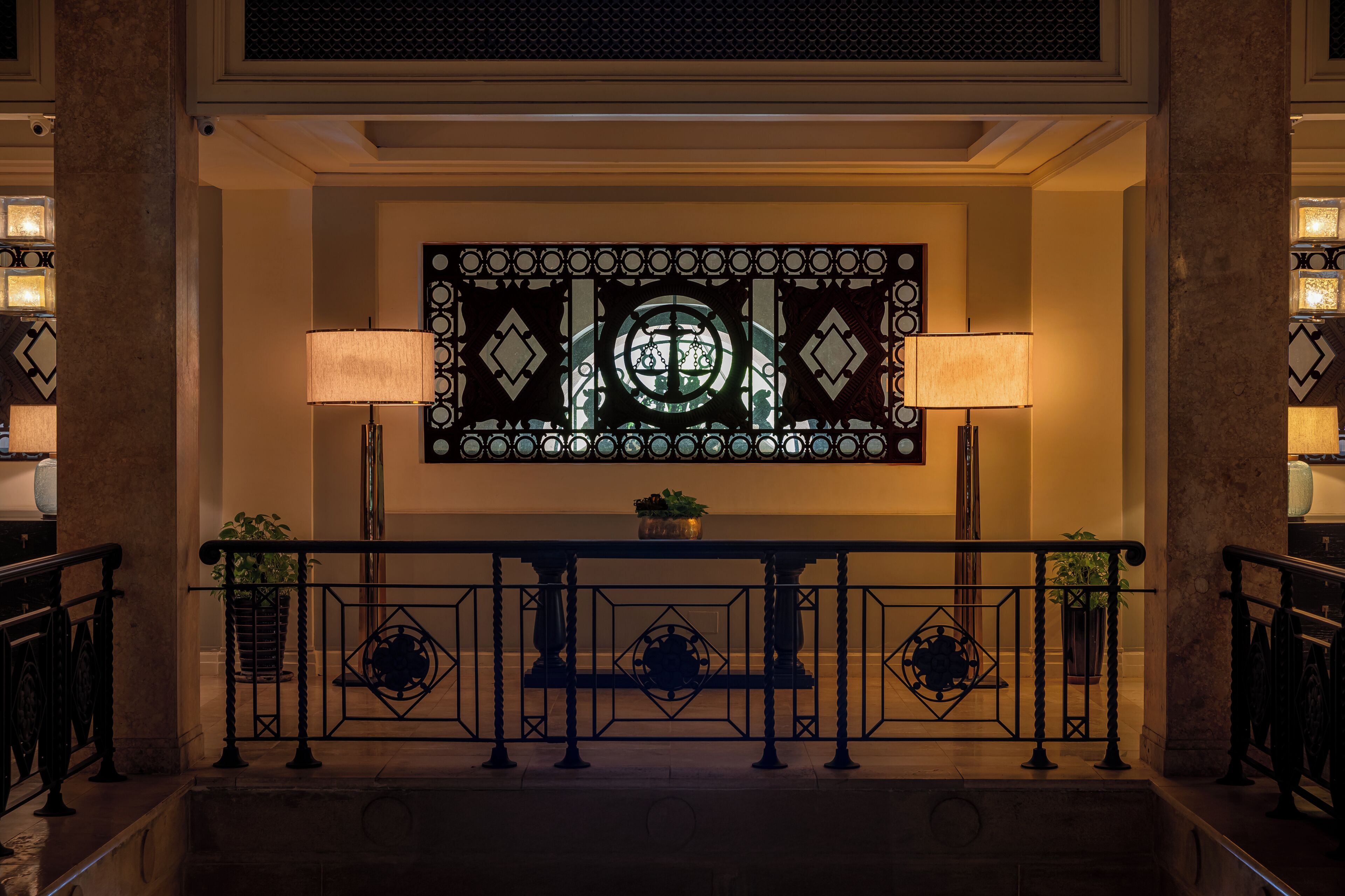 Un vestíbulo imponente con un ventanal decorativo de vitral, una mesa central con planta en maceta y una barandilla de metal negro.