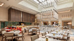 Un vestíbulo de hotel espacioso con varias zonas de restauración, un bar central y un alto techo con una lámpara de araña moderna.