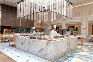 Un amplio vestíbulo de hotel con un mostrador de recepción de mármol, un chef preparando comida y una lámpara de araña moderna.
