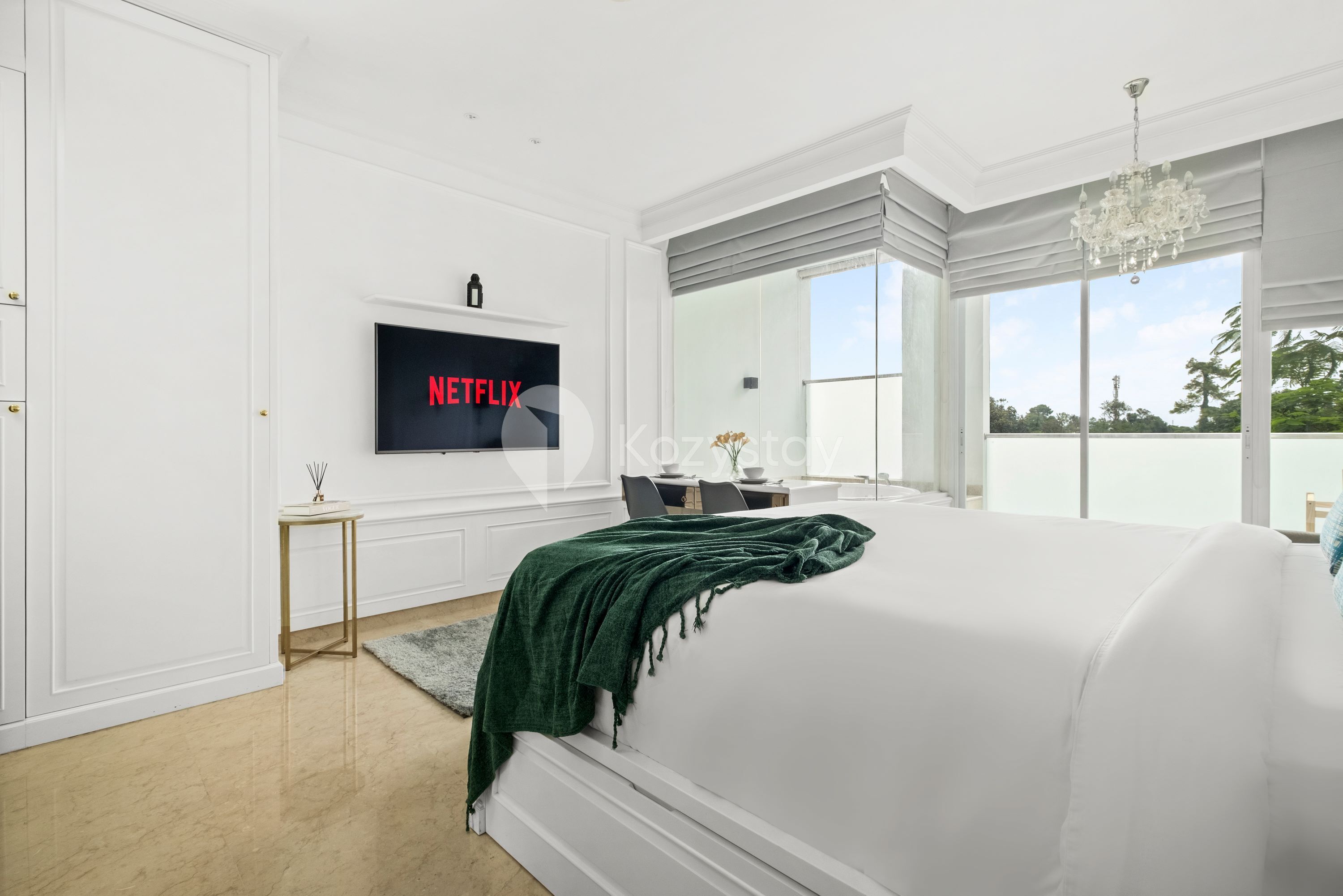 Ein Schlafzimmer mit einem großen Bett, einem an der Wand befestigten Fernseher, einem Schreibtisch mit Stuhl und Blick ins Freie durch große Fenster.