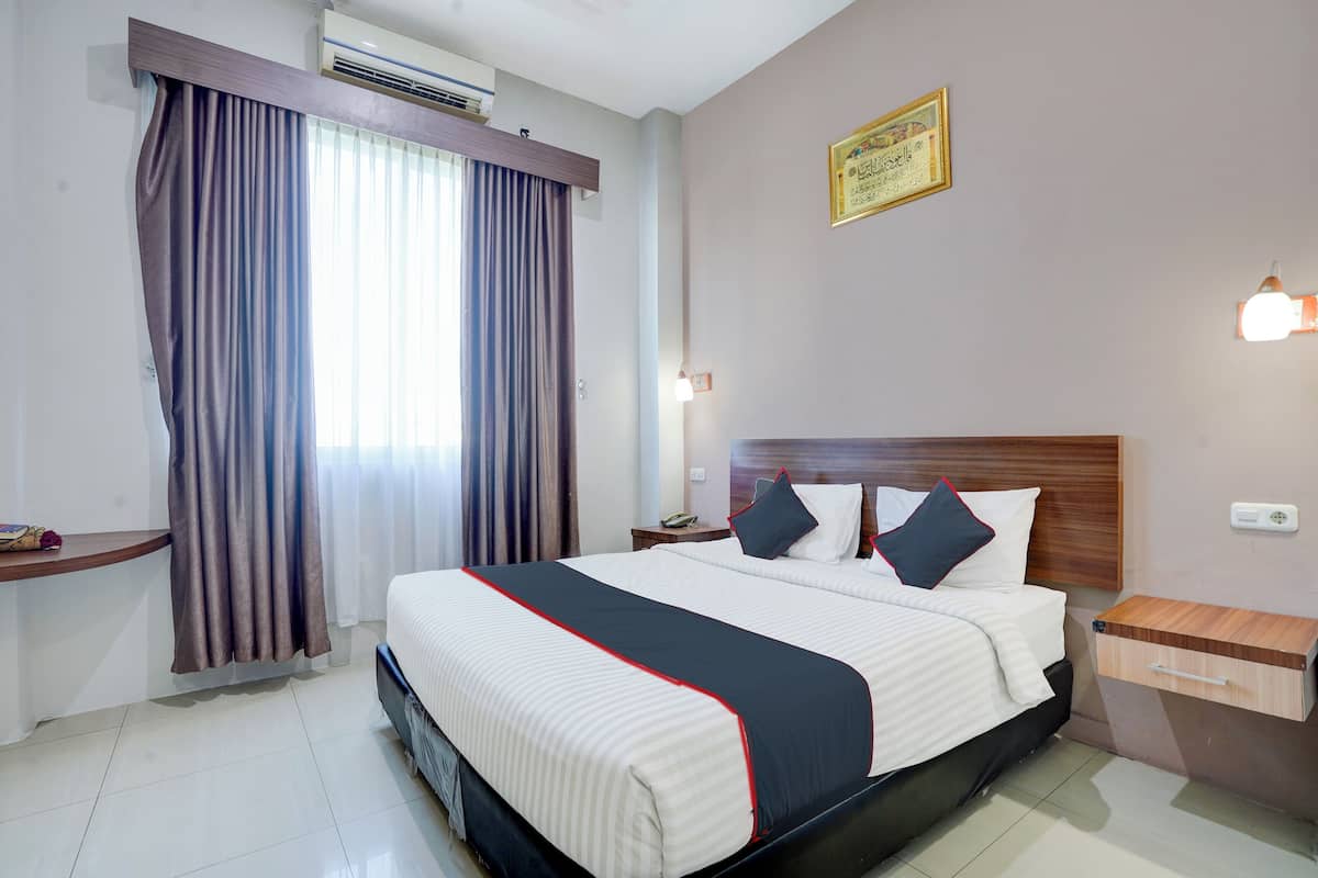 Une chambre d’hôtel avec un grand lit, une table de chevet, une commode, une étagère fixée au mur et une fenêtre avec des rideaux.