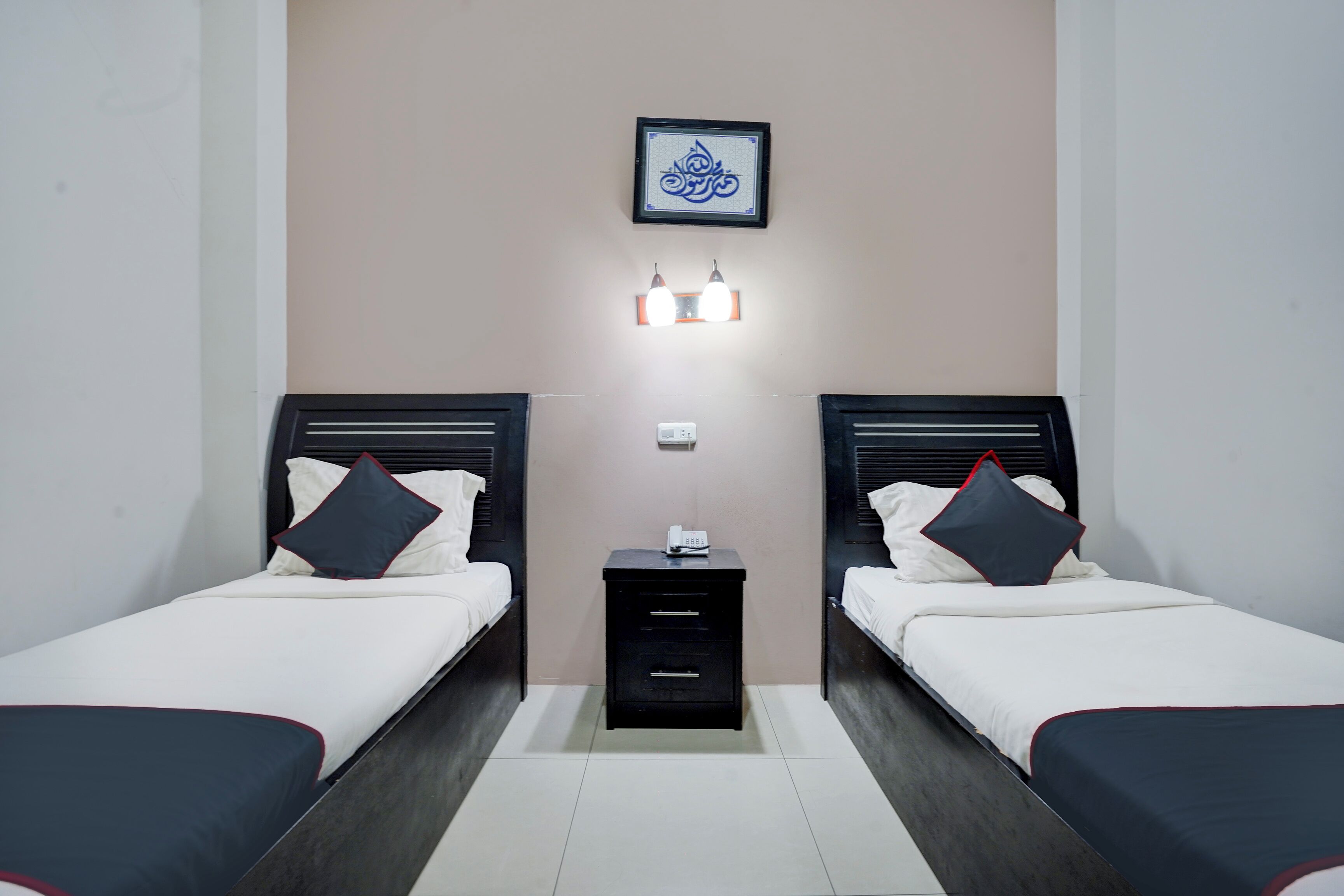 Quarto de hotel com duas camas, uma mesa de cabeceira, luminária fixada na parede e uma obra de arte emoldurada sobre a mesa.