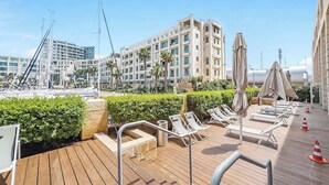 A rooftop deck with sun loungers, umbrellas, and a view of a marina and buildings.