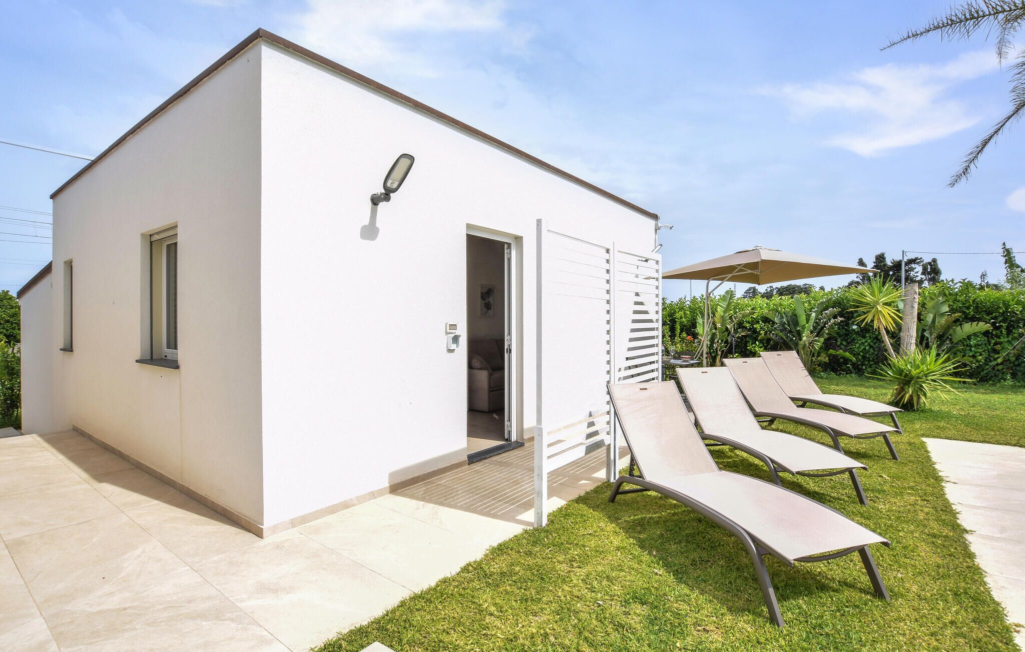 Un bâtiment moderne, de plain-pied, avec une façade blanche, une porte et une petite fenêtre. On aperçoit en arrière-plan deux fauteuils de jardin, une table et un parasol.