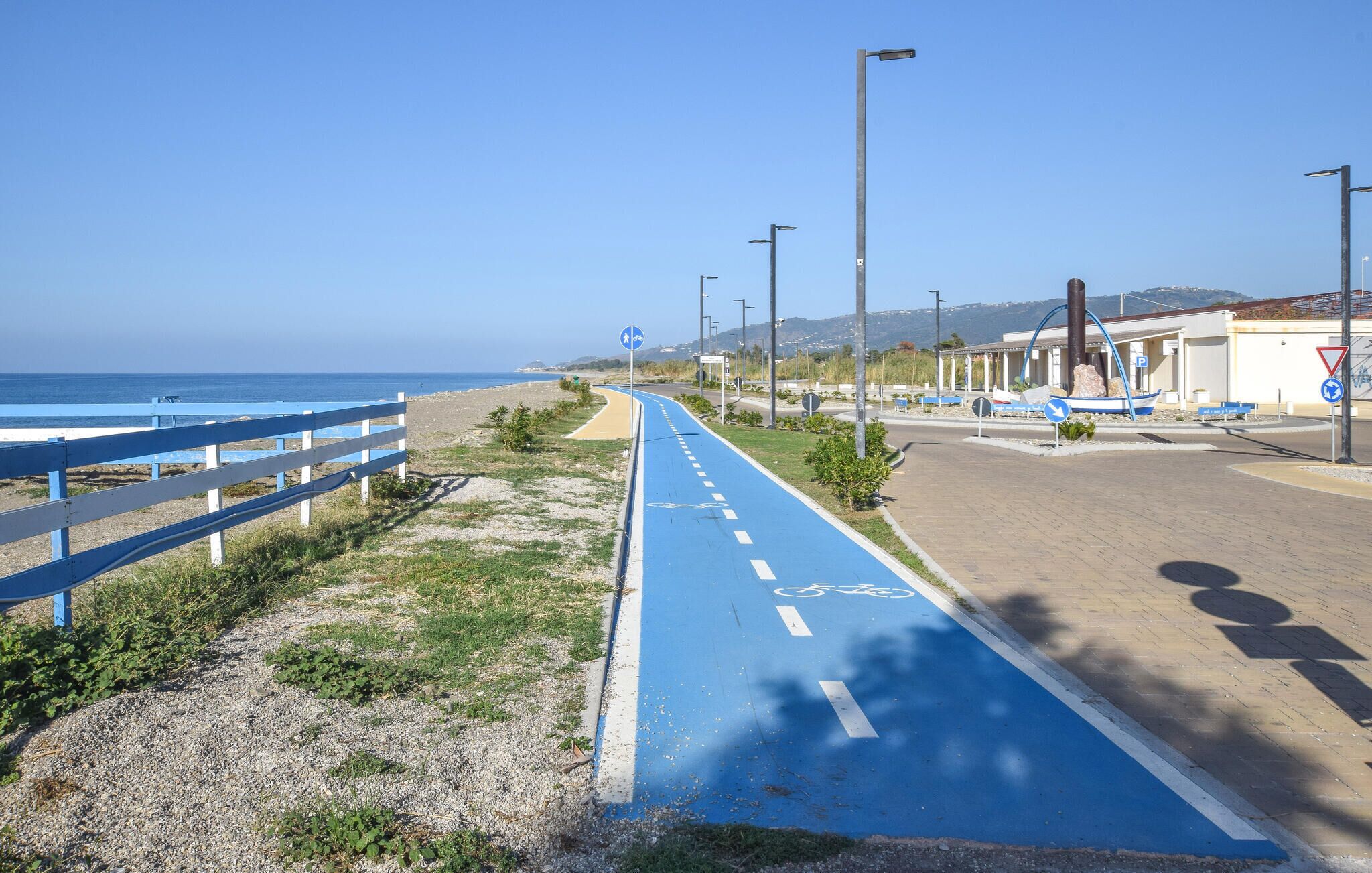 Eine Küstenstraße mit blau markierter Radwegspur, Fußweg und Strand.