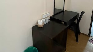 Una habitación pequeña con un escritorio de madera oscura, un espejo, un cubo de basura verde y una pared blanca con toma de corriente.