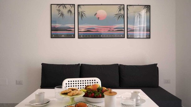 A dining table with a meal setup, including pastries, fruit, and a glass of water, in front of a sofa with a cityscape painting above it. - Casa Marisa, Salento by design. Within walking distance of the pearls of Salento! (Torre Saracena)
