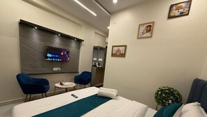 A hotel room with a bed, two chairs, a small table, a TV, and a wall-mounted shelf. - SHARAN'S INN SUTES & BANQUETS (Hyderabad)
