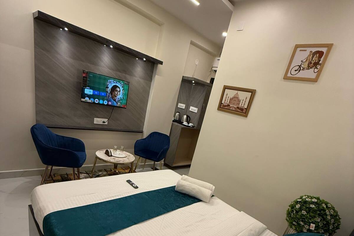 Una habitación de hotel moderna con una cama grande, dos sillas azules, una mesita redonda y un televisor montado en la pared.