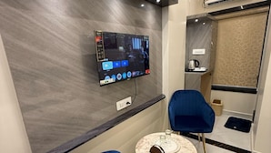 A modern room with a flat-screen TV mounted on a dark wall, two blue chairs, a small round table, and a coffee machine. - SHARAN'S INN SUTES & BANQUETS (Hyderabad)