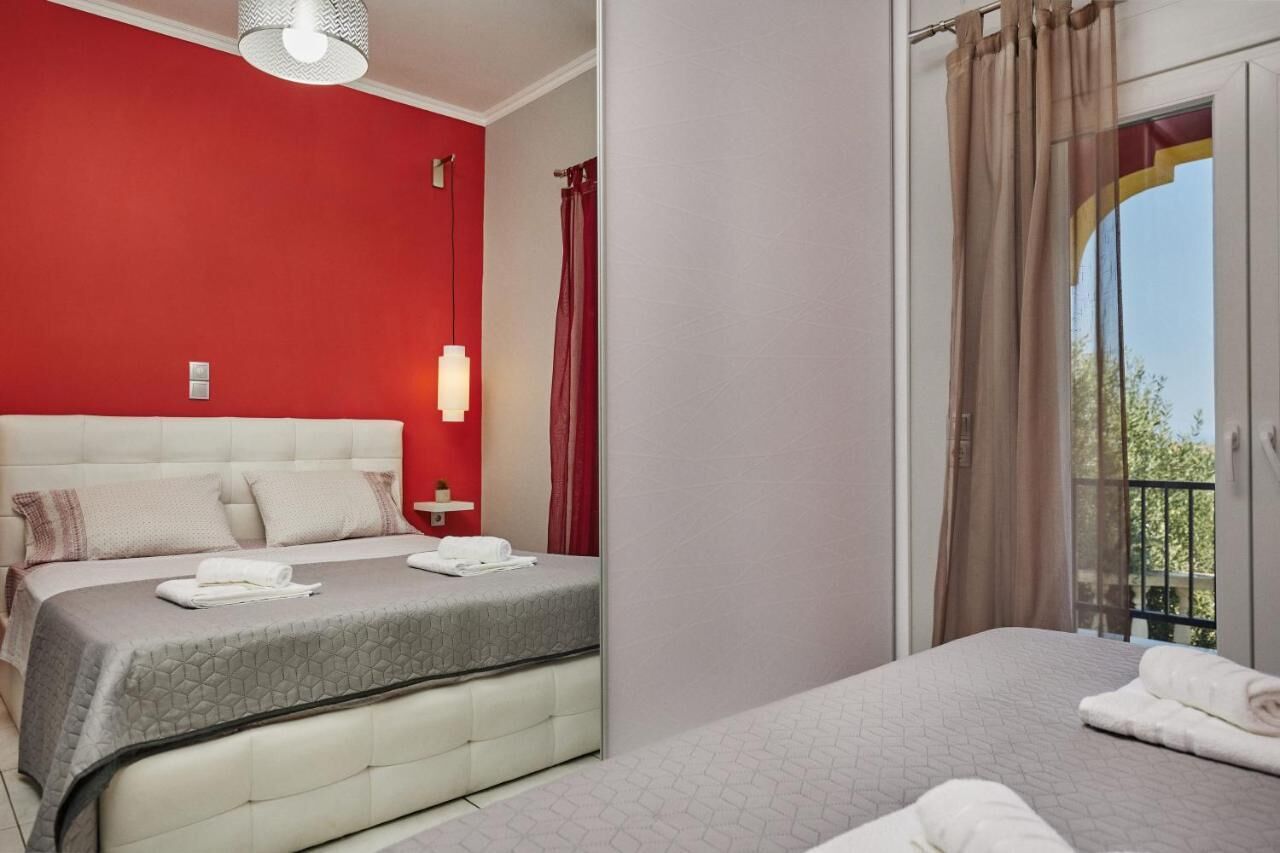 Hotelkamer met een rode accentmuur, een groot bed met een grijze quilt, een wit hoofdeinde en een balkon met gordijnen.