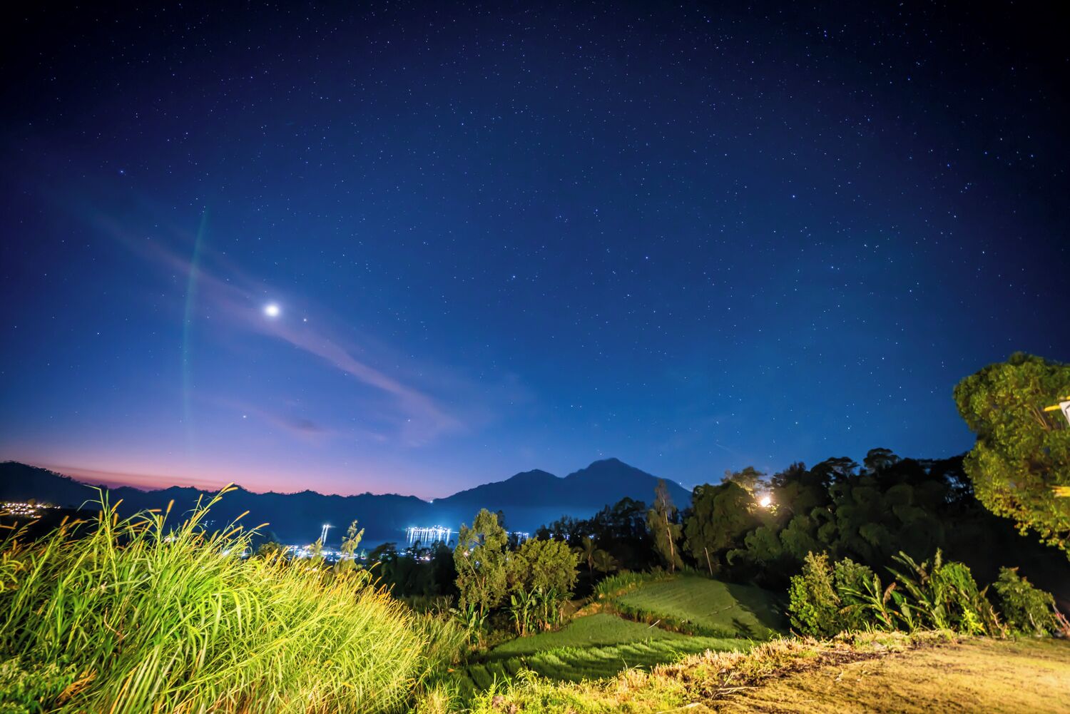 Cielo nocturno con estrellas, una cadena montañosa y un edificio iluminado en la distancia.
