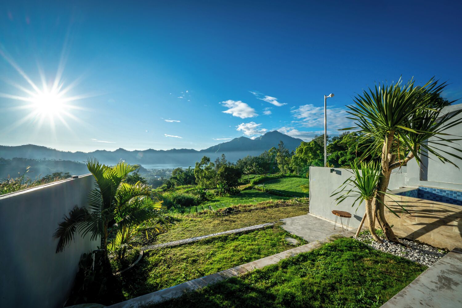 A residential area with a view of mountains and a body of water. There is a concrete pathway leading to a patio area with a chair and a palm tree.