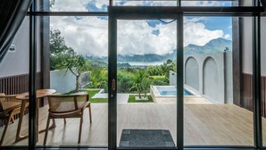 Habitación de hotel moderna con un gran ventanal corredero que da a un balcón con piscina, mobiliario exterior y vistas a montañas y vegetación.