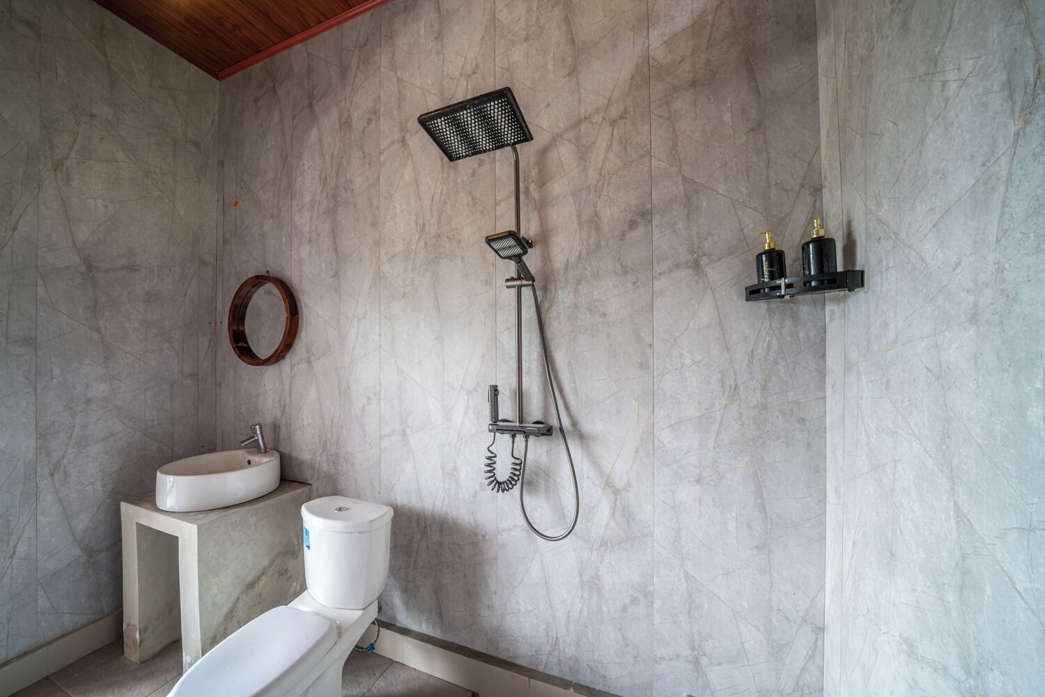 Ein Badezimmer mit Marmorwand, einem weißen Waschbecken, einer Toilette, einer Dusche inklusive Handbrause und einem kleinen Ablagebereich mit Flaschen.