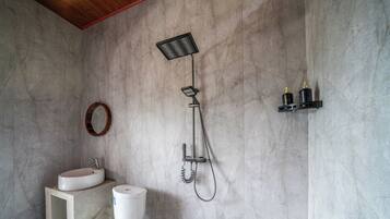 Ein Badezimmer mit Marmorwand, einem weißen Waschbecken, einer Toilette, einer Dusche inklusive Handbrause und einem kleinen Ablagebereich mit Flaschen.