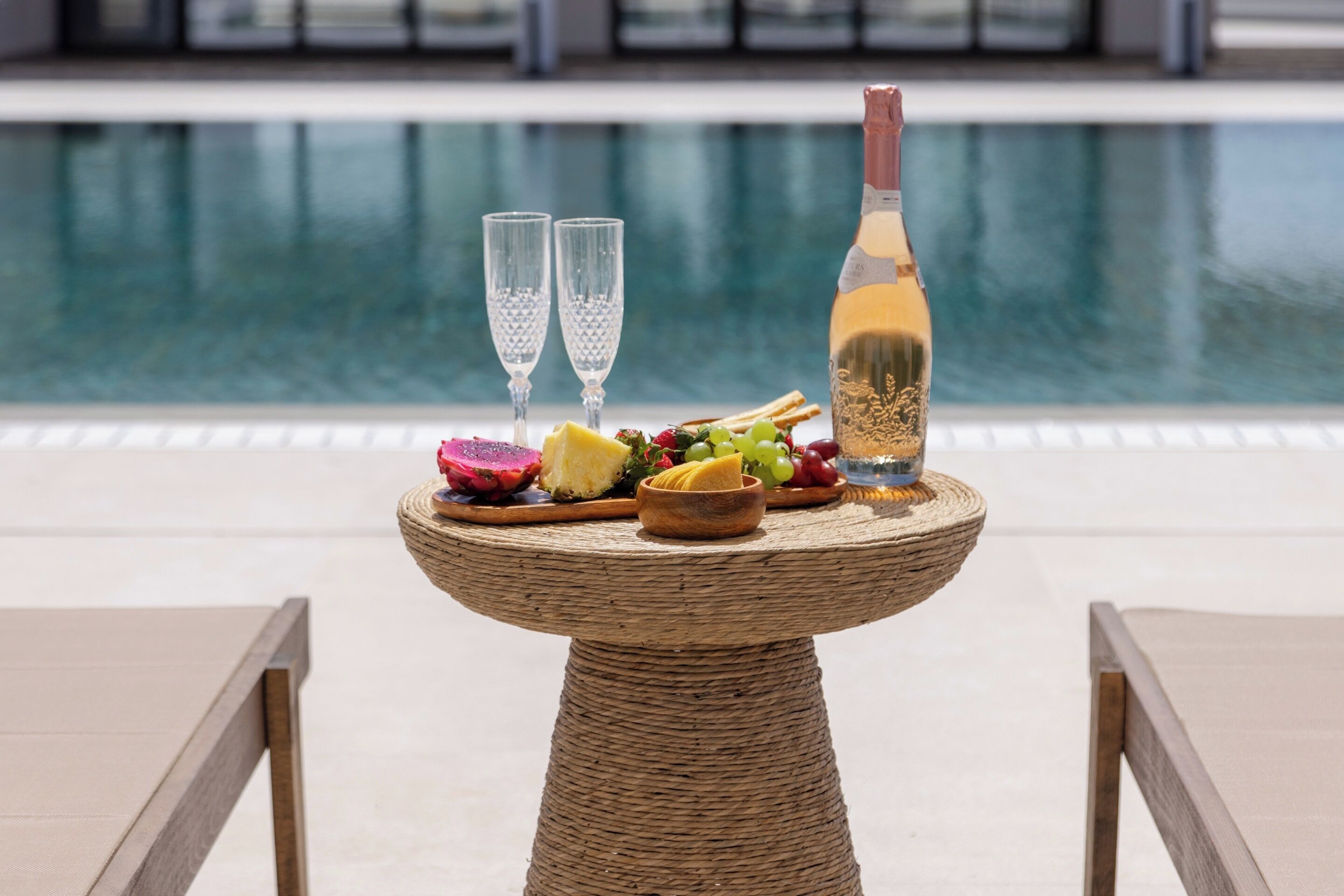 A wicker table with a bottle of white wine, two glasses, and a variety of fruits and cheeses on a terrace by a pool.