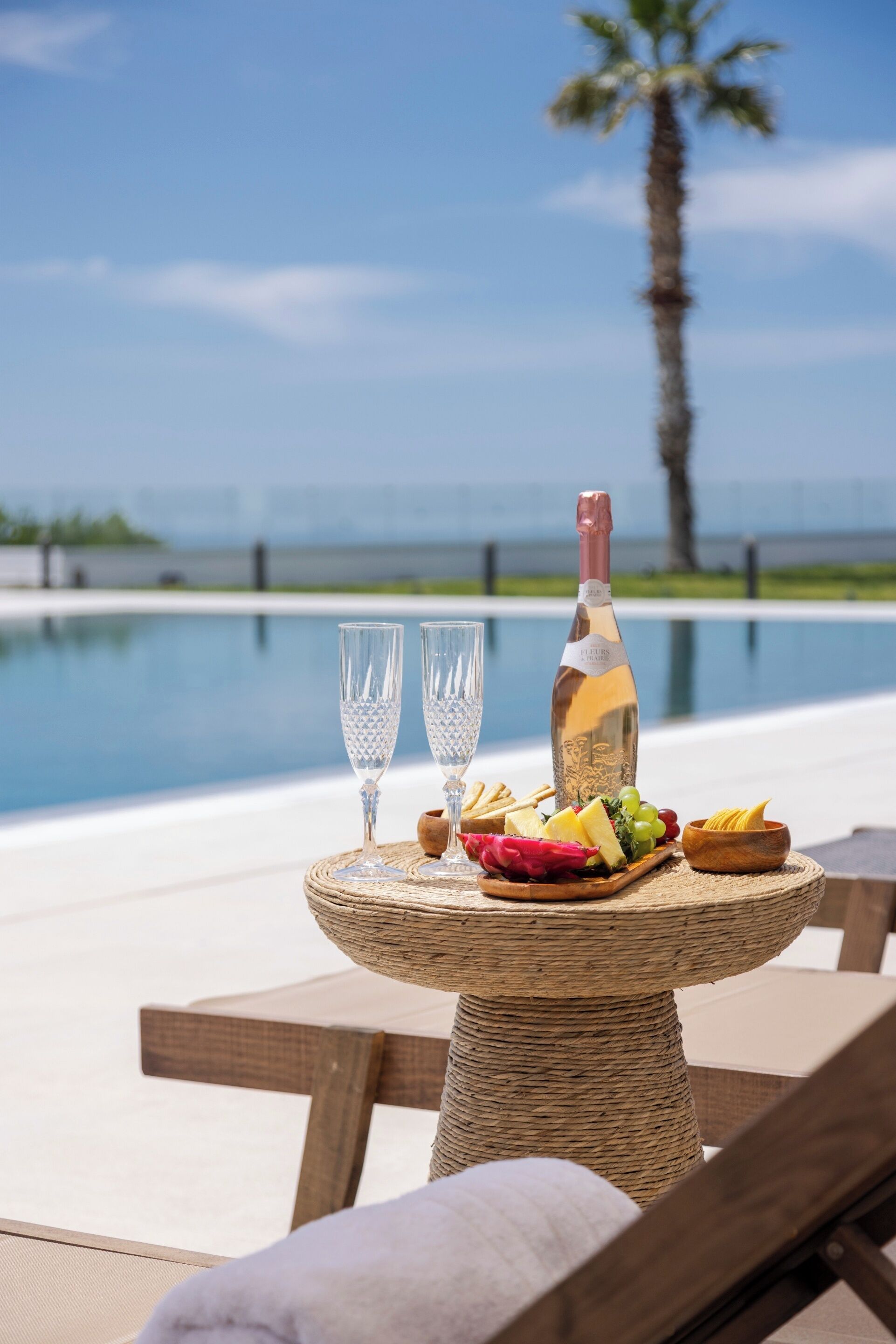 A table with a bottle, glasses, and a fruit platter by a pool with a palm tree in the background.