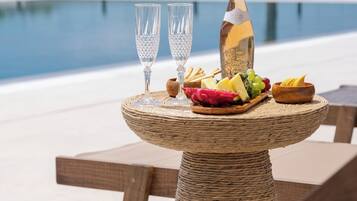A table with a bottle, glasses, and a fruit platter by a pool with a palm tree in the background.