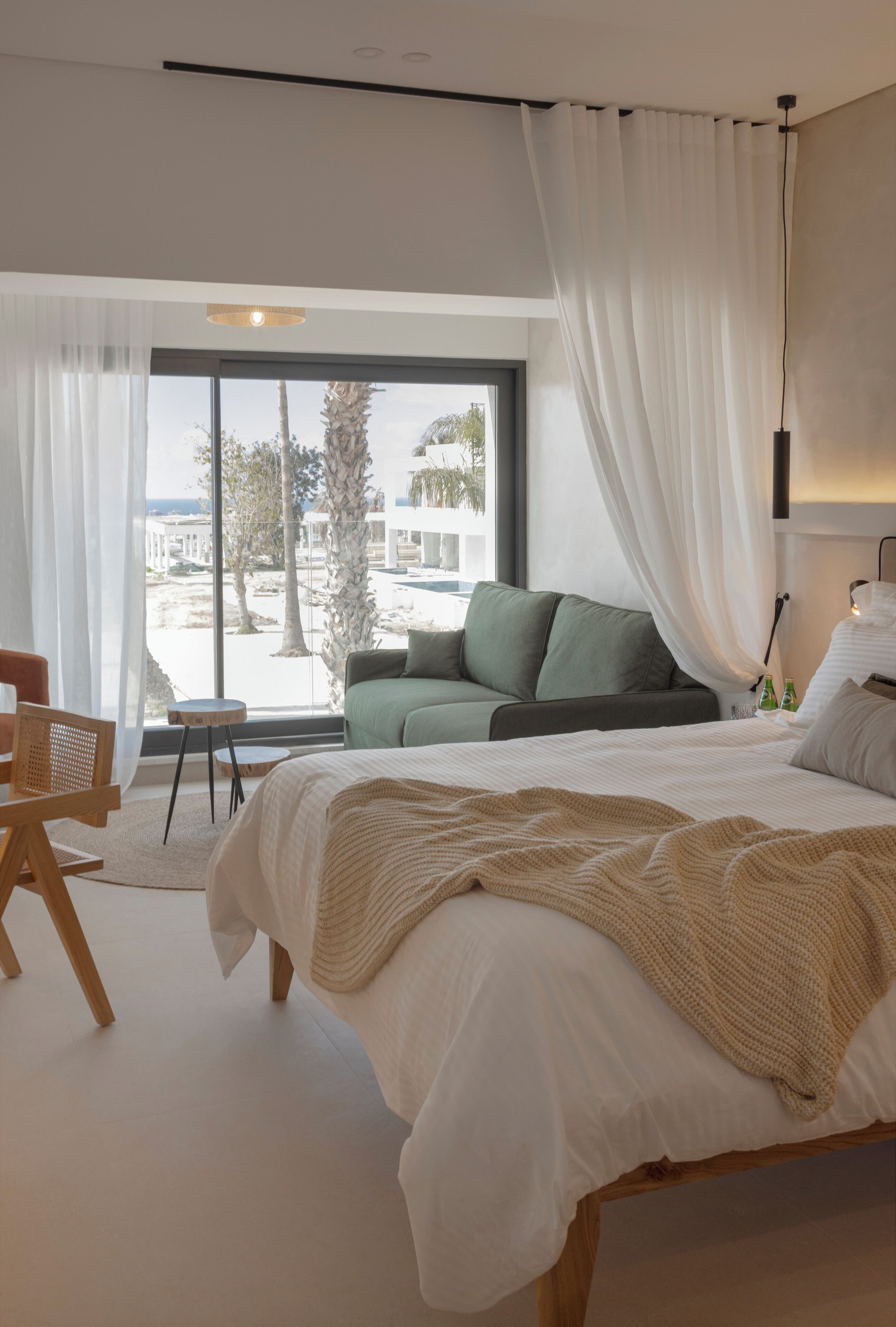 A modern bedroom with a large bed, a green sofa, and a view of palm trees outside.