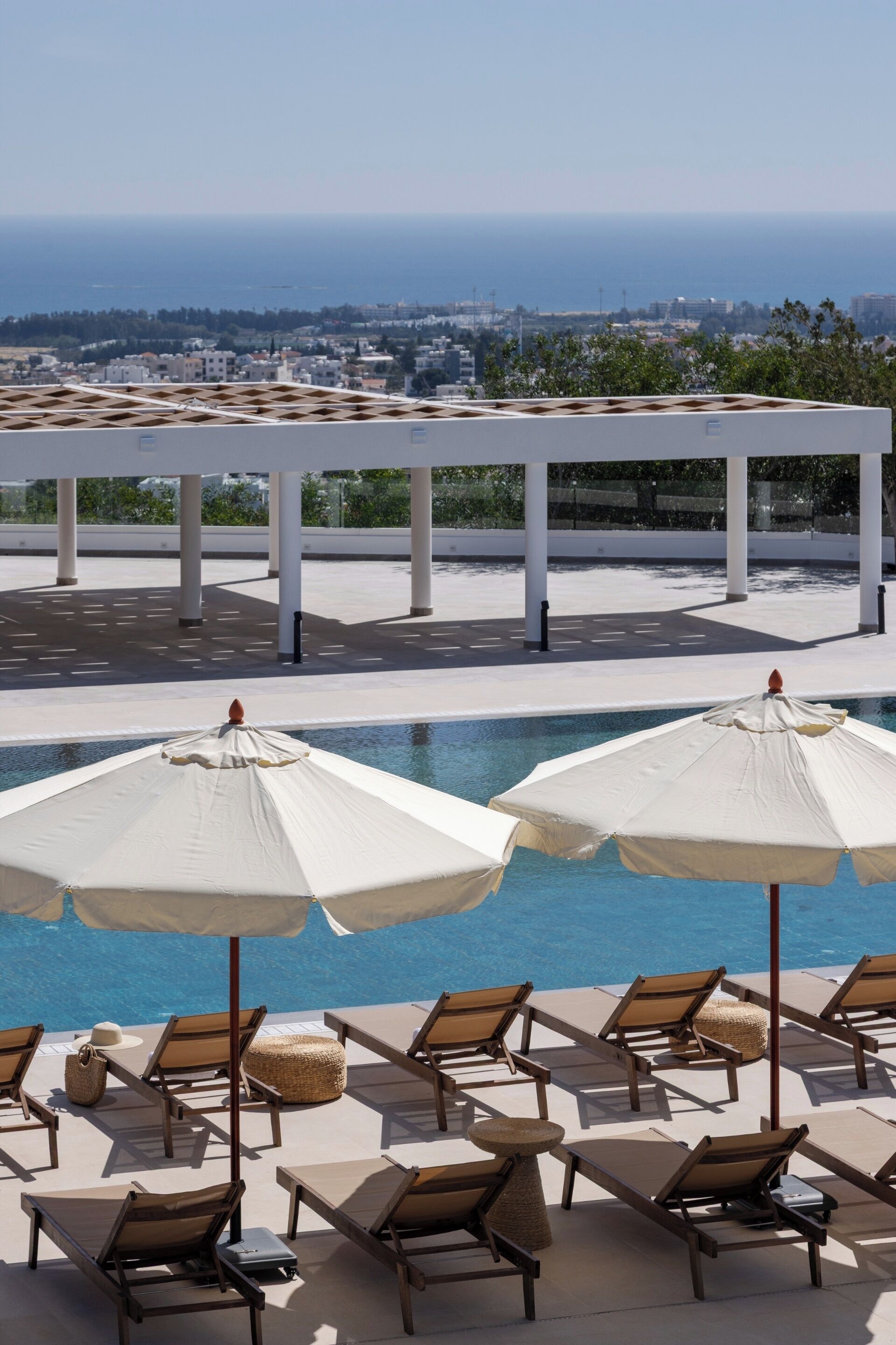A poolside area with lounge chairs and umbrellas, a clear view of the sea, and a white pergola structure.