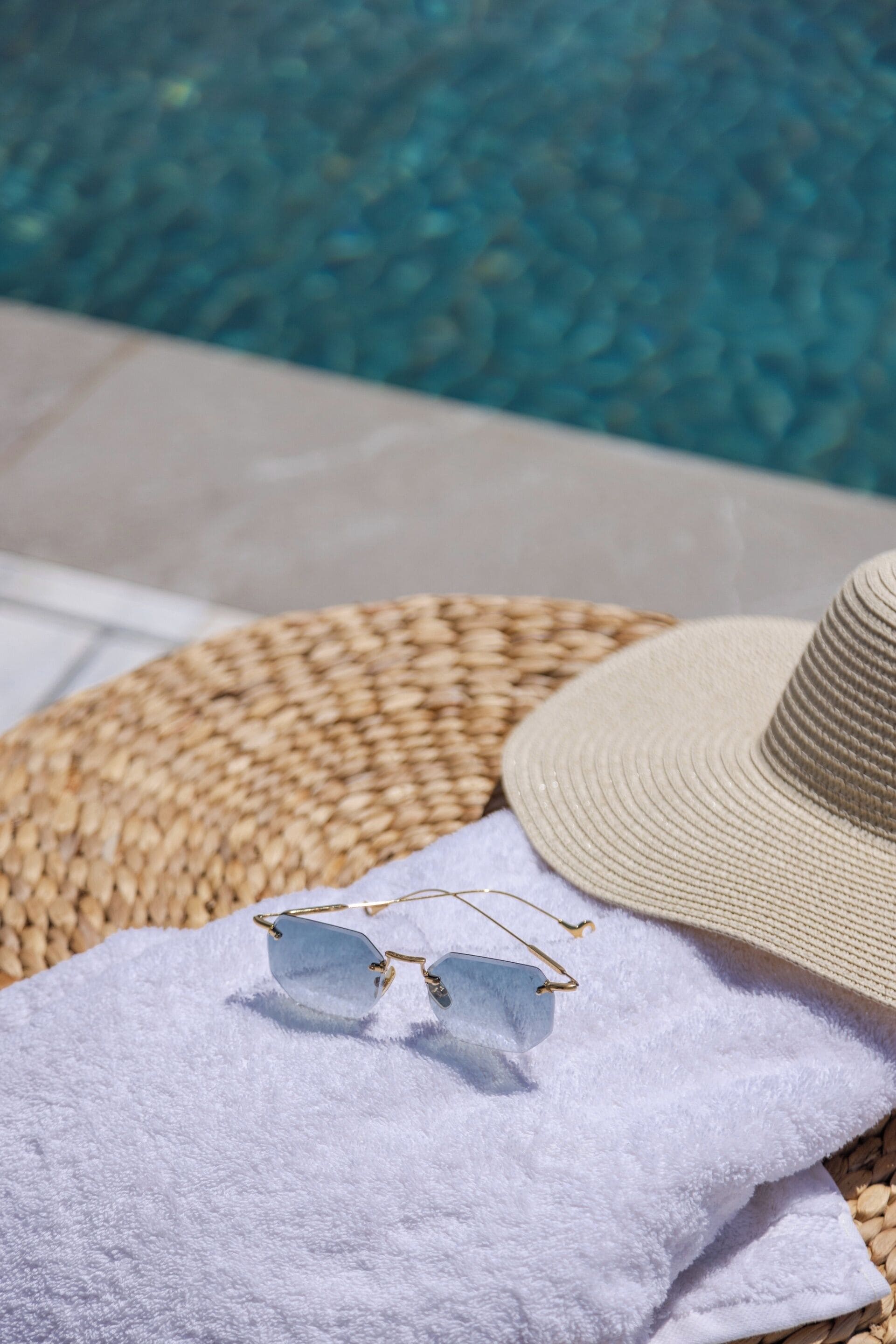 Una estera trenzada, un sombrero de paja y unas gafas de sol sobre una toalla blanca junto a una piscina.