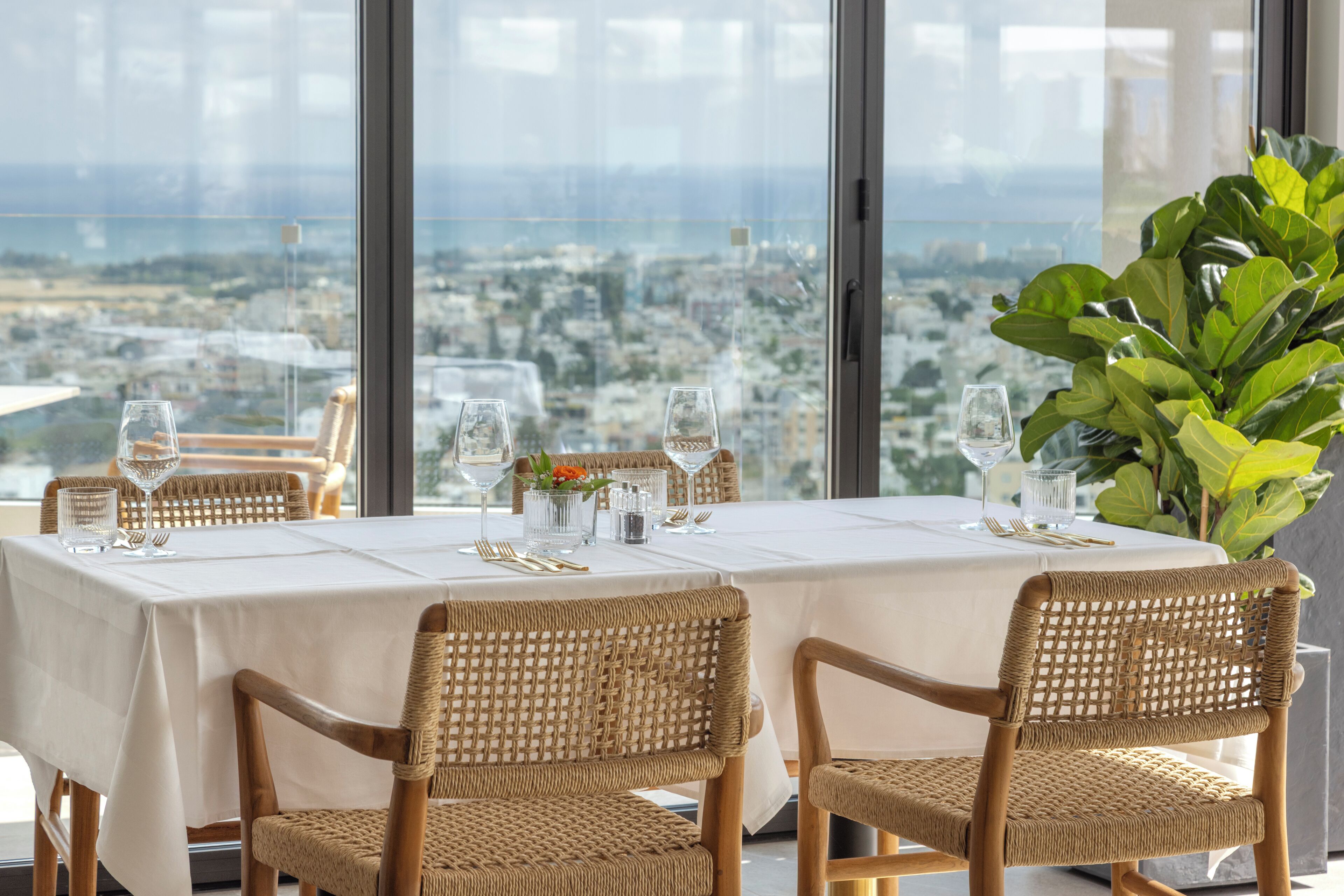 Mesa de comedor con mantel blanco, cristalería y sillas de mimbre, con vistas a un paisaje urbano.