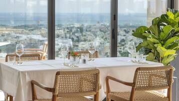 Mesa de comedor con mantel blanco, cristalería y sillas de mimbre, con vistas a un paisaje urbano.