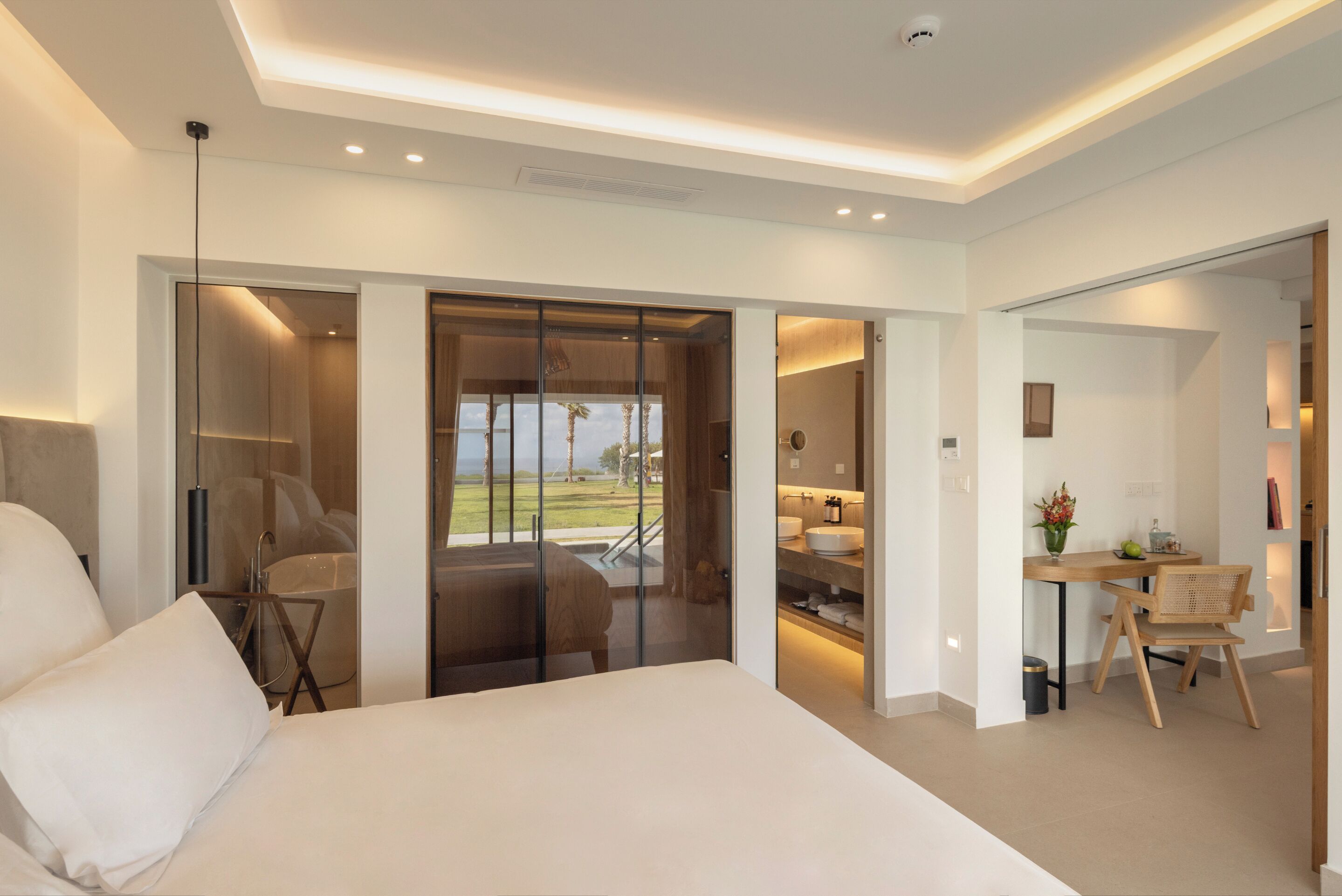 A modern hotel room with a large bed, a desk with a chair, a bathroom with a glass door, and a view of the outdoors.