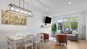 A dining area with a white table and chairs, a modern pendant light, a flat-screen TV on a shelf, and a large window with a view of greenery.