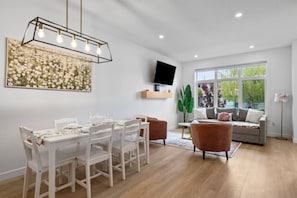 Un comedor con una mesa y sillas blancas, una lámpara colgante moderna, un televisor de pantalla plana sobre una estantería y un ventanal con vistas a la vegetación.