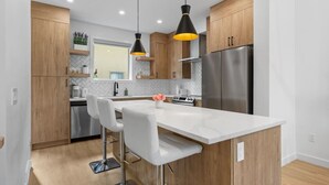 A modern kitchen with wooden cabinets, a stainless steel refrigerator, a white marble island, and pendant lighting.