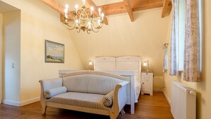 A bedroom with a wooden floor, a striped sofa, a white bed, a nightstand, a chandelier, and a radiator. - Charming semi-detached house in Trent on Rügen - Landhaus Trent Haushälfte Breet (Trent)